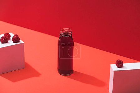 fresh berry juice in glass bottle near ripe raspberries on cubes on red background